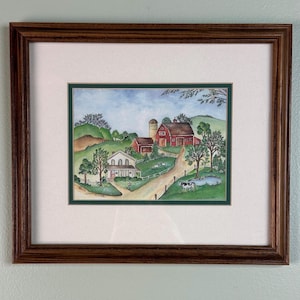 May include: Framed watercolor painting of a rural farm scene. The artwork depicts a white house, red barns, a silo, and a winding road. The painting is set in a green landscape with trees, a pond, and cows. The frame is a dark brown wood.