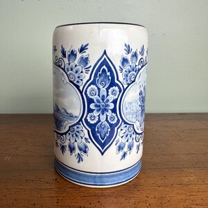 Op de afbeelding: Cilindrische keramische vaas met een witte basis en blauwe bloemmotieven. De vaas heeft een centraal blauw bloemmotief en twee landschapssc&egrave;nes. De boven- en onderranden zijn afgewerkt met blauwe banden. De vaas is ongeveer 15 cm hoog.