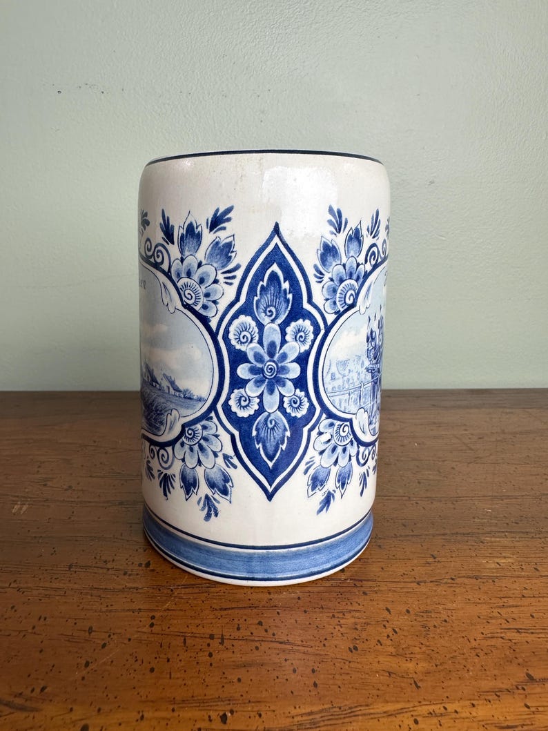Op de afbeelding: Cilindrische keramische vaas met een witte basis en blauwe bloemmotieven. De vaas heeft een centraal blauw bloemmotief en twee landschapssc&egrave;nes. De boven- en onderranden zijn afgewerkt met blauwe banden. De vaas is ongeveer 15 cm hoog.