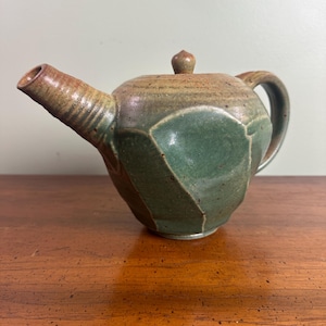 May include: Handmade ceramic teapot with a green and brown glaze. The teapot features a unique geometric design on the body, a curved handle, and a spout. The lid has a small knob. The teapot is a functional piece of art.