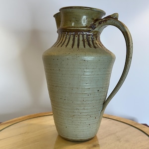 May include: A stoneware pitcher with a speckled light green glaze and a brown rim. The body features horizontal ridges and a series of vertical brown markings around the neck. It has a single curved handle and a wide, flared pouring spout.