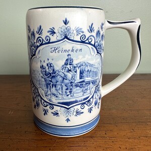 Op de afbeelding: Een witte keramische bierpul met blauwe bloemmotieven en het woord "Heineken". De pul heeft een gedetailleerde illustratie van een paardenkoets. De handgreep is wit met een blauwe rand.