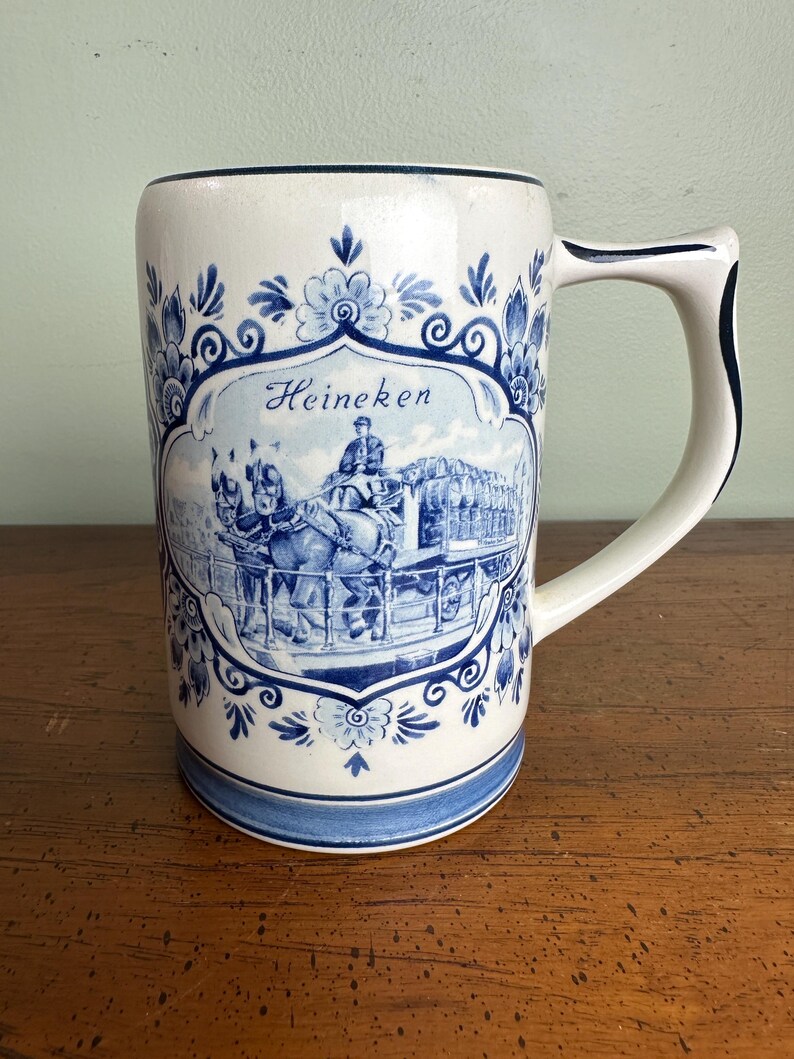 Op de afbeelding: Een witte keramische bierpul met blauwe bloemmotieven en het woord "Heineken". De pul heeft een gedetailleerde illustratie van een paardenkoets. De handgreep is wit met een blauwe rand.