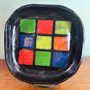 May include: A square black ceramic dish with a colorful mosaic tile design. The mosaic features nine squares in red, green, blue, and yellow. The dish has a slightly irregular shape and a glossy finish.