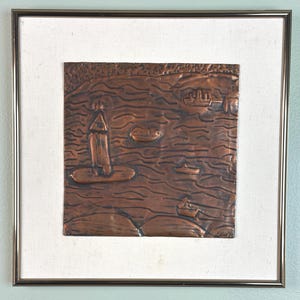 May include: A copper relief artwork depicting a coastal scene with a lighthouse, boats, and water. The artwork is framed with a silver metal frame and mounted on a white canvas. The copper has a rich, warm brown color.