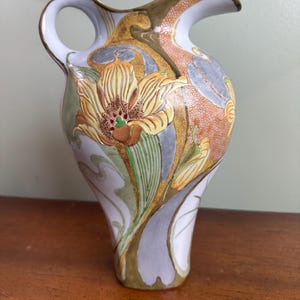 May include: A decorative ceramic pitcher with a white base, featuring a floral design in shades of yellow, brown, and green. The pitcher has a curved handle and spout, with gold accents throughout the design. The pitcher is approximately 10 inches tall.