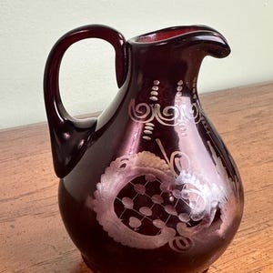 May include: A deep red glass pitcher with a curved handle and a spout. The pitcher features etched floral and scroll designs in a lighter shade of red. The pitcher is likely used for serving beverages and adds a touch of elegance to any table setting.