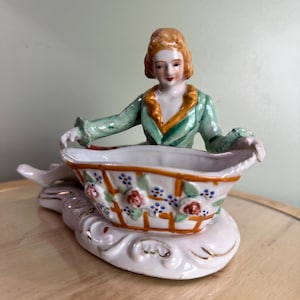 May include: A vintage porcelain figurine of a woman in a green jacket with an orange collar, holding a basket. The basket is white with a floral design of red roses and blue flowers. The figurine is on a white base with gold trim.