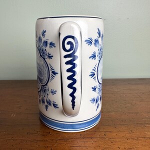 Op de afbeelding: Witte keramische mok met blauwe bloemen- en architectuurpatronen. De mok heeft een blauw handvat met een spiraalvormig ontwerp en een blauwe band aan de basis. Het ontwerp doet denken aan Delfts blauw.