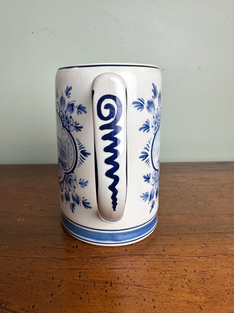 Op de afbeelding: Witte keramische mok met blauwe bloemen- en architectuurpatronen. De mok heeft een blauw handvat met een spiraalvormig ontwerp en een blauwe band aan de basis. Het ontwerp doet denken aan Delfts blauw.