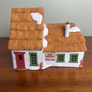 May include: A ceramic cottage figurine with a thatched roof, snow accents, and green window frames. The red door and chimney add color. The sign reads "Bob Cratchit Tiny Tim."
