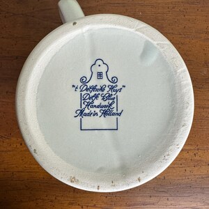 Op de afbeelding: Onderkant van een cr&egrave;mekleurige keramische mok met een handvat. De basis toont blauwe tekst en een huisillustratie, met de tekst "'t Delftsche Huys' Delft Blue Handwork Made in Holland."