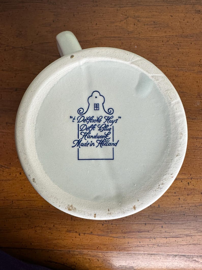 Op de afbeelding: Onderkant van een cr&egrave;mekleurige keramische mok met een handvat. De basis toont blauwe tekst en een huisillustratie, met de tekst "'t Delftsche Huys' Delft Blue Handwork Made in Holland."