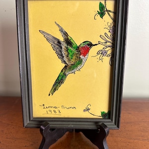 May include: Framed artwork featuring a hummingbird in flight, with green, red, and white plumage, feeding from purple flowers. The artwork is set against a yellow background and framed in a dark gray frame. Signed "Leona Brunner 1983".