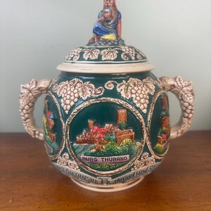 May include: A decorative ceramic stein with a lid featuring a seated figure. The dark green body has relief carvings of grapes and three circular scenes with castles. The stein has two handles and the text "Burg Thurand" is visible.