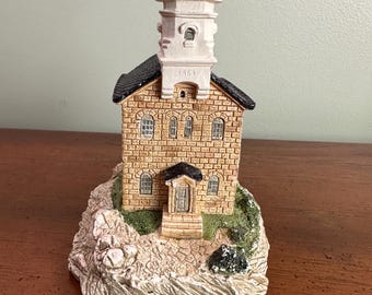 May include: A detailed miniature lighthouse figurine. The lighthouse features a brick-like structure with a black roof, white tower, and a red top. The base is a rocky terrain with green accents. The year "1863" is visible on the tower.
