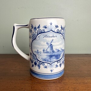 Op de afbeelding: Een witte keramische bierpul met blauwe bloemen- en windmolenontwerpen. De pul heeft een handvat en een rechthoekige vorm. Het woord "Heineken" staat in script op de voorkant. Het ontwerp doet denken aan Delfts blauw.