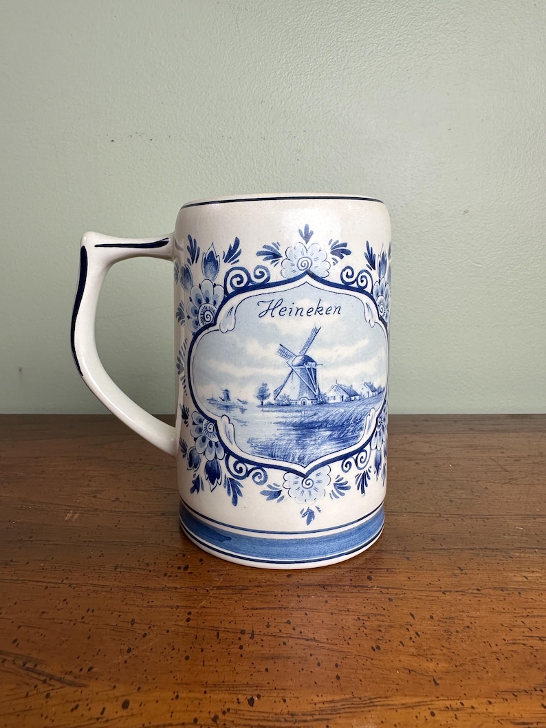 Op de afbeelding: Een witte keramische bierpul met blauwe bloemen- en windmolenontwerpen. De pul heeft een handvat en een rechthoekige vorm. Het woord "Heineken" staat in script op de voorkant. Het ontwerp doet denken aan Delfts blauw.