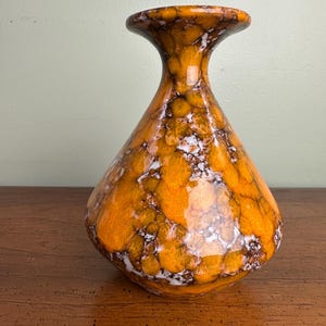 May include: A decorative ceramic vase with a bulbous body and flared rim. The vase features a glossy orange glaze with a mottled pattern of brown and white, creating a unique, textured appearance. The vase is a decorative home accent.