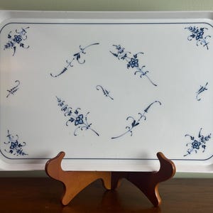 May include: A rectangular white serving tray with a raised edge, adorned with blue floral patterns. The tray is supported by a wooden stand. The floral design is symmetrical, with delicate blue flowers and leaves.
