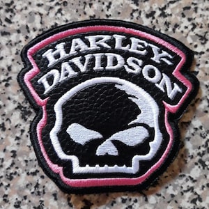Op de afbeelding: Een zwarte geborduurde patch met een witte schedelafbeelding en de tekst Harley Davidson in witte letters. De patch heeft een zwarte achtergrond met textuur en is omlijnd met een felroze rand, wat zorgt voor een opvallend contrast.