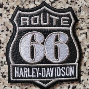 Puede incluir: Parche negro y gris de Route 66 Harley-Davidson. El parche presenta las palabras "ROUTE 66" en la parte superior, el número "66" en el centro y "HARLEY-DAVIDSON" en la parte inferior. El parche tiene forma de escudo con un borde negro.