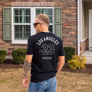 Puede incluir: Camiseta negra con el texto blanco "LOS ANGELES 23 BASKETBALL". La camiseta la lleva una persona con gafas de sol y vaqueros azules. La persona está de pie frente a una casa con revestimiento de ladrillo.