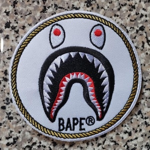 May include: A circular white patch featuring a shark face design with red eyes, a black mouth, and white teeth. The patch is bordered by a gold rope trim and includes the text "BAPE®" in black.