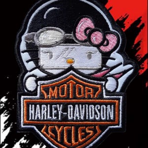 Peut inclure: Un écusson brodé représentant un personnage de chat blanc portant un casque noir avec des lunettes et un nœud rose, placé au-dessus d'un logo en forme de bouclier Harley-Davidson Motor Cycles noir et orange avec du texte blanc.