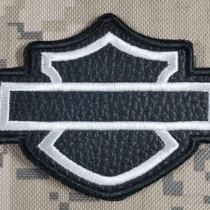 Puede incluir: Un parche bordado en blanco y negro con un emblema en forma de escudo y una barra horizontal en el centro. El parche tiene una superficie negra texturizada con un borde blanco cosido que delinea el escudo y la barra central.
