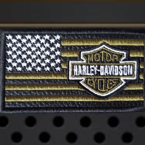 May include: A rectangular embroidered patch featuring a stylized American flag design with black and gold horizontal stripes and a white star field. The Harley-Davidson Motor Cycles shield logo is embroidered in white and gold over the stripes.