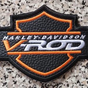 Peut inclure: Écusson Harley-Davidson V-Rod noir et orange. De forme de bouclier, il présente un fond noir et des bordures orange. Le texte "HARLEY-DAVIDSON" est arqué au-dessus de "V ROD", écrit dans une police audacieuse et stylisée.