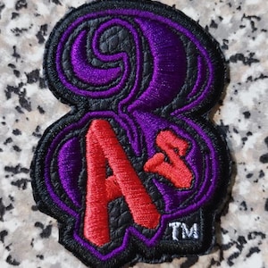 Puede incluir: Un parche bordado en negro, morado y rojo que presenta el número 8 estilizado con las letras A y s integradas en el diseño. El parche tiene un borde negro e incluye las letras TM en hilo blanco en la esquina inferior derecha.