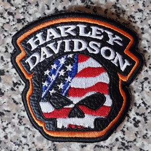 Puede incluir: Un parche bordado en forma de escudo con el texto Harley Davidson en letras blancas sobre un fondo negro. El centro muestra un gráfico de calavera relleno con un patrón de bandera estadounidense roja, blanca y azul, bordeado por un ribete naranja.