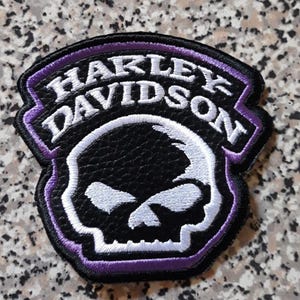 May include: A black and purple embroidered patch featuring a white skull graphic and the text Harley Davidson in white lettering. The patch has a textured black background and a purple border, with a shield-like shape and white stitching.