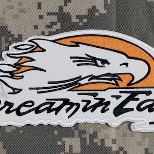 Puede incluir: Parche bordado con la cabeza de un águila blanca con detalles en naranja, sobre un círculo naranja. Las palabras "Screamin' Eagle" están escritas debajo en una fuente estilizada. El parche tiene un borde blanco.
