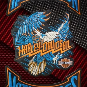 Puede incluir: Parches bordados de Harley-Davidson con un diseño de águila azul y naranja y el nombre de la marca. Los parches están sobre un fondo texturizado negro y rojo. También se muestra la palabra "Motorcycles".