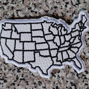 Puede incluir: Un parche de piel sintética blanca con la forma del mapa de los Estados Unidos. El parche presenta contornos bordados en negro que definen las fronteras de cada estado. Los bordes del parche están rematados con un borde bordado en blanco.