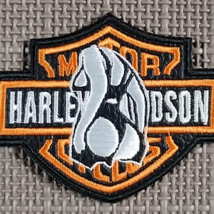 Może przedstawiać: Haftowana naszywka z logo Harley-Davidson w kolorach czarnym, pomarańczowym i białym. Naszywka ma kształt tarczy z napisem "HARLEY DAVIDSON" i "MOTOR" w białych literach.
