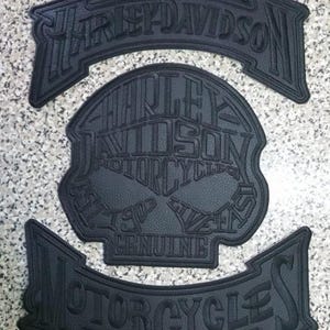 Może przedstawiać: Trzy czarne naszywki motocyklowe Harley-Davidson. Górna i dolna naszywka są łukowate z logo Harley-Davidson. Środkowa naszywka ma kształt czaszki z logo Harley-Davidson i napisem "GENUINE".