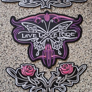 Puede incluir: Colección de parches bordados con temática motera. El parche más grande presenta un diseño de mariposa con las palabras "LIVE LOVE RIDE". Otros parches incluyen remolinos decorativos y rosas rosas. Los parches son negros, blancos, plateados y morados.