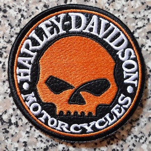Puede incluir: Un parche bordado circular que presenta una calavera naranja central con cuencas oculares negras. La calavera está rodeada por un borde negro con el texto blanco HARLEY-DAVIDSON MOTORCYCLES. El diseño está enmarcado por finos anillos cosidos en naranja y negro.