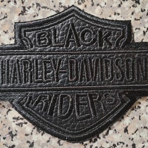 Op de afbeelding: Zwarte Harley-Davidson Riders patch met geborduurde tekst. De patch heeft de vorm van een schild met een rechthoekig midden, met het Harley-Davidson logo en de woorden "BLACK RIDERS". De patch is gemaakt van textuurmateriaal.