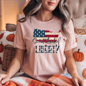Puede incluir: Camiseta rosa claro con un gráfico del mapa de Estados Unidos con el diseño de la bandera americana. El texto "Sweet Land of Liberty" está impreso debajo del mapa en una fuente de escritura y letras en bloque.