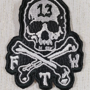 Könnte beinhalten: Schwarz-weißer, gestickter Aufnäher mit einem Totenkopf mit der Zahl 13 darüber. Unter dem Schädel befinden sich gekreuzte Knochen, und die Buchstaben F, T und W sind sichtbar.