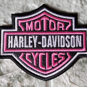 Puede incluir: Un parche bordado en forma de escudo con el logotipo de Harley-Davidson. El diseño incluye hilo negro, blanco y rosa. El texto dice MOTOR, HARLEY-DAVIDSON y CYCLES en letras blancas y rosas sobre un fondo negro.