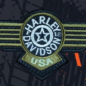 May include: A black and olive green embroidered patch featuring a central circular emblem with a star and the text Harley-Davidson. The design includes stylised wings extending horizontally and a bottom tab with the text USA in olive green.