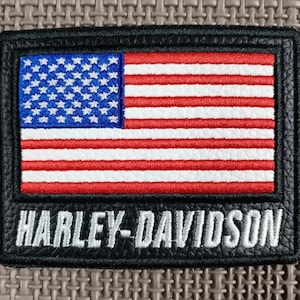 Puede incluir: Un parche rectangular negro con una bandera estadounidense bordada en rojo, blanco y azul. El texto "HARLEY-DAVIDSON" está bordado en blanco debajo. El parche tiene un borde cosido.