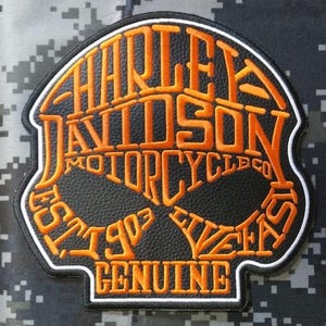 Op de afbeelding: Een zwarte patch met een getextureerd oppervlak en oranje geborduurde tekst die HARLEY DAVIDSON MOTORCYCLE CO EST 1903 LIVE FAST GENUINE leest. De patch heeft een witte en zwarte rand en is gevormd als een gestileerd schedelsilhouet.