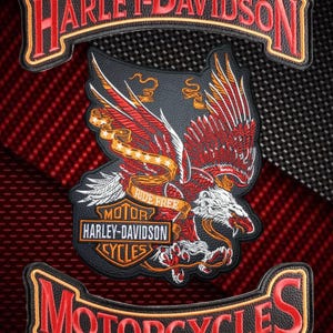 Puede incluir: Un conjunto de tres parches bordados que incluye un logo de Harley-Davidson en negro y rojo en la parte superior y uno de Motorcycles en la inferior. El parche central muestra un águila detallada con alas rojas y una cinta dorada con Ride Free.